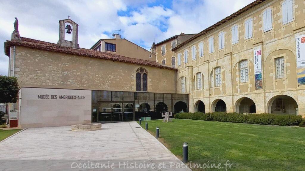 L'entrée moderne en verre du Musée des Amériques-Auch s'harmonise avec les bâtiments historiques en pierre beige ; des banderoles sont accrochées à la façade, avec une petite statue sur le toit à gauche, une pelouse et une allée soigneusement entretenues au premier plan.