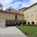 L'entrée moderne en verre du Musée des Amériques-Auch s'harmonise avec les bâtiments historiques en pierre beige ; des banderoles sont accrochées à la façade, avec une petite statue sur le toit à gauche, une pelouse et une allée soigneusement entretenues au premier plan.