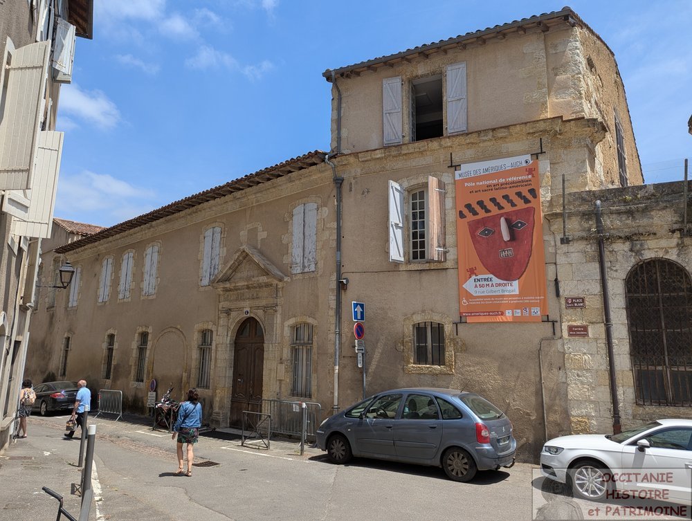 Vue de la rue de l'ancienne entrée du musée avec ses bâtiments en pierre beige orné d'une grande affiche artistique orange, avec des passants à proximité et des voitures garées le long du trottoir sous un ciel bleu et ensoleillé.