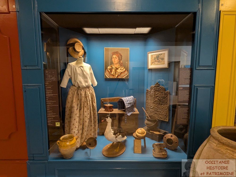 Vitrine de musée présentant des vêtements traditionnels, un portrait peint, un canard empaillé et divers outils ménagers historiques.