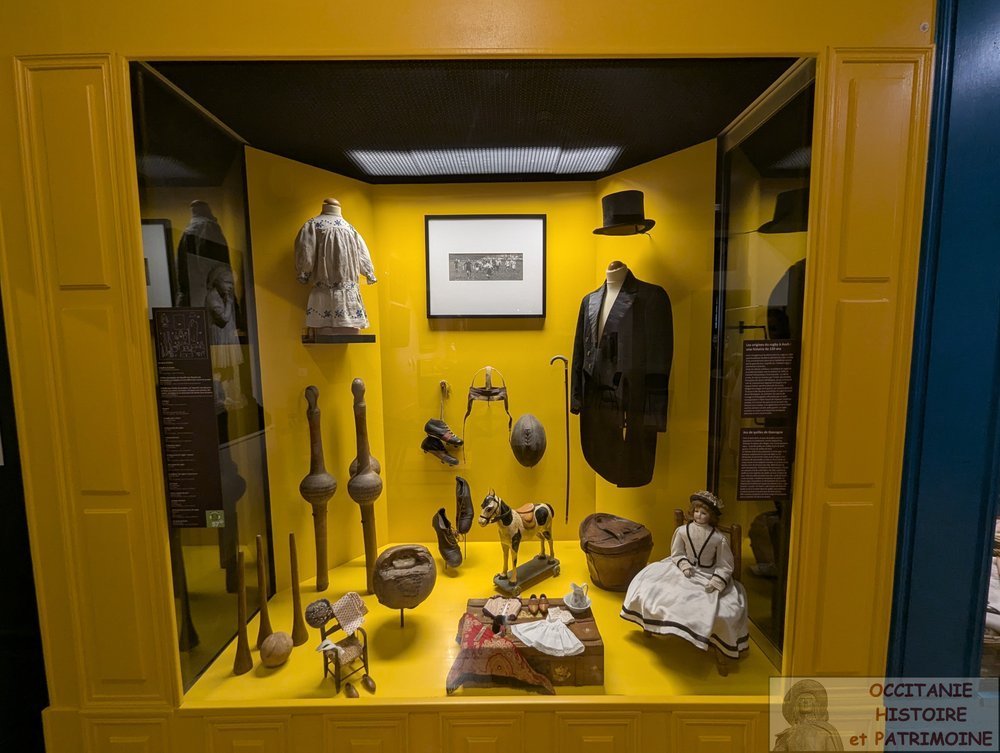 Une vitrine de musée jaune contenant divers vêtements d'époque, des objets en bois dont un ancien jeu de quille, un cheval en bois, une poupée ainsi que de vieux crampons et un ballon de rugby.