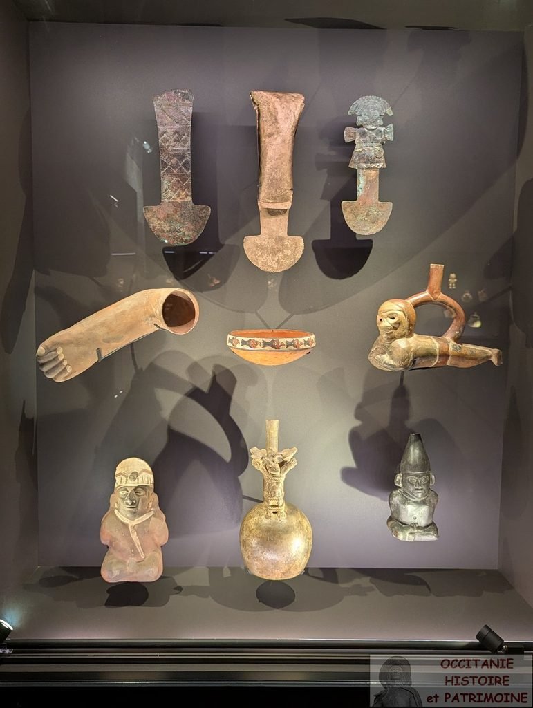 Une vitrine de musée contenant sept objets rituels, notamment des outils, des poteries et des figurines, disposés sur deux étagères devant un fond sombre.