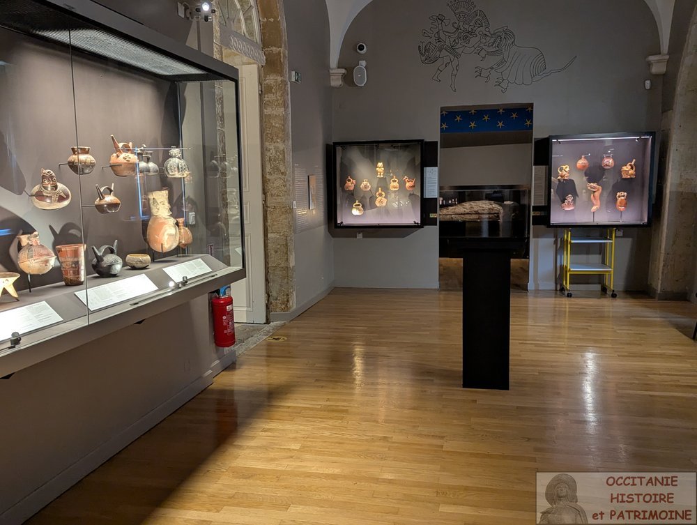 Une exposition muséale présente des poteries amérindiennes dans des vitrines éclairées, accompagnées de panneaux explicatifs et d'un extincteur fixé au mur. Le sol est en bois et la salle est dotée de piliers en pierre voûtés.