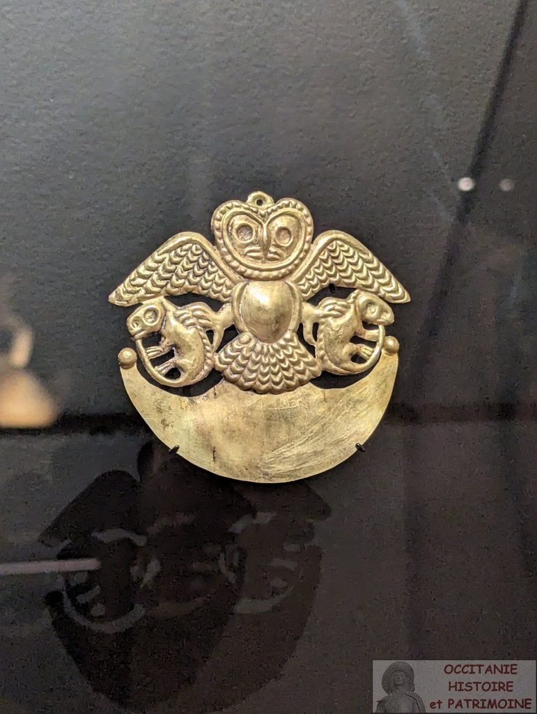 Broche en or en forme de chouette aux ailes déployées, perchée sur un croissant de lune, flanquée de deux créatures ressemblant à des lézards. Présentée sur un fond sombre.