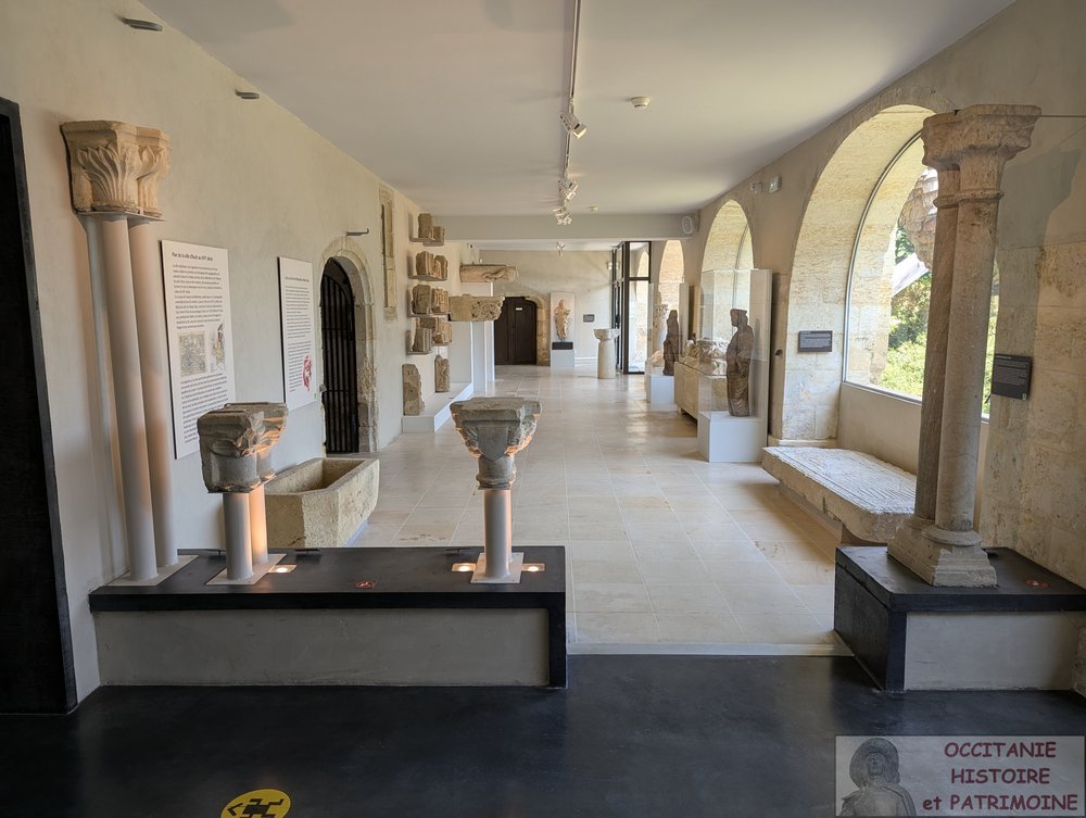 Vaste pièce en longueur du musée avec des colonnes en pierre, des sculptures et des objets anciens qui y sont exposés. Des murs blancs et de grandes fenêtres cintrées laissent entrer la lumière naturelle. Des panneaux explicatifs et des socles sont visibles un peu partout dans l'espace.