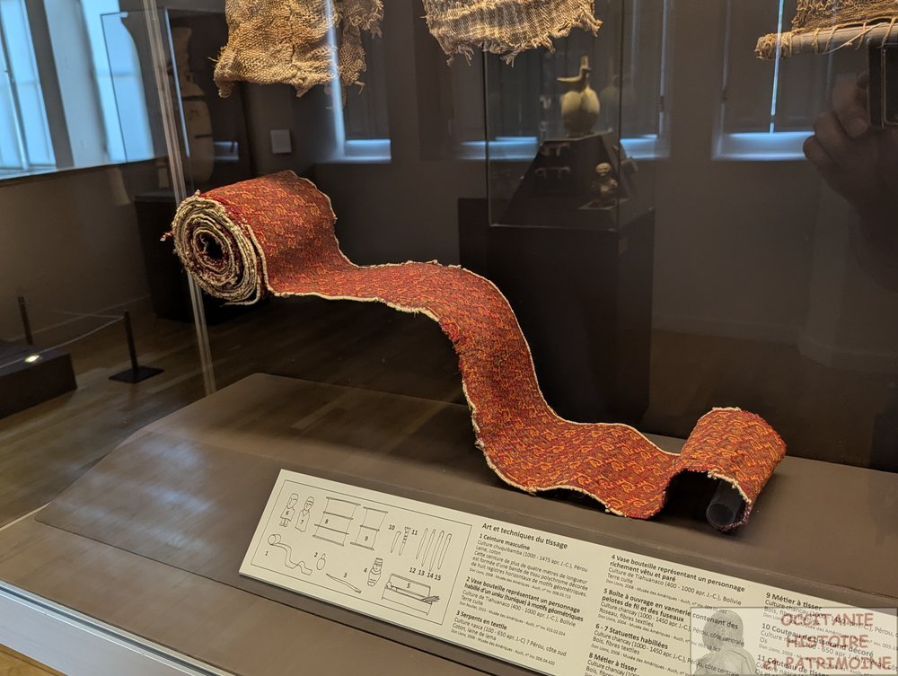 Un long tissu enroulé, orné d'un motif rouge et or, est exposé dans une vitrine, accompagné d'une plaque explicative comportant des schémas et un texte en français. D'autres objets et morceaux de tissu sont visibles en partie au dessus.