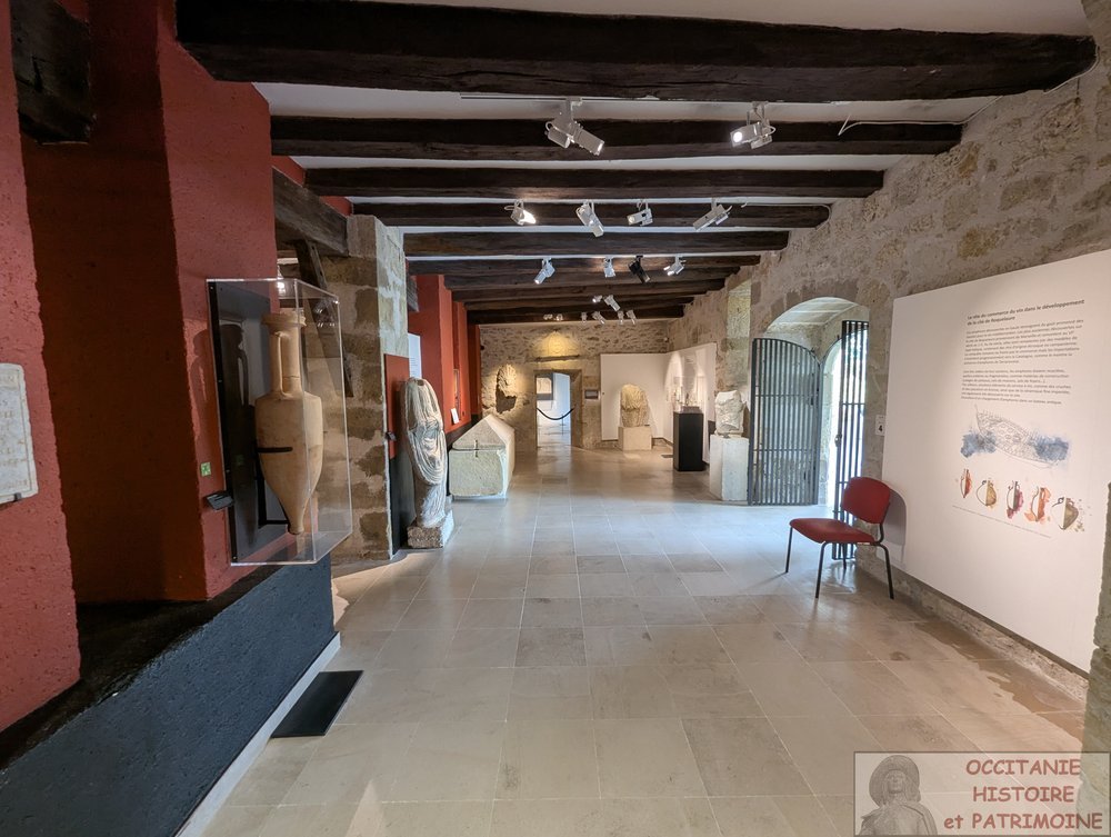 Une vaste pièce en longueur de musée aux murs en pierre, où l'on peut voir des objets antiques, des vitrines, des panneaux explicatifs et une chaise rouge. Un éclairage sur rail met en valeur les objets exposés et des poutres en bois ornent le plafond.
