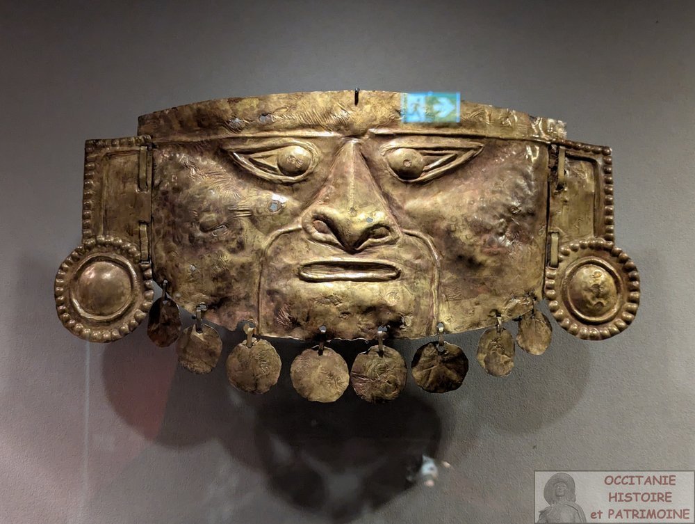 Masque amérindien en or, au visage sévère, aux grands yeux et au nez proéminent ; des ornements circulaires pendent en dessous. Le masque est exposé sur un fond gris.