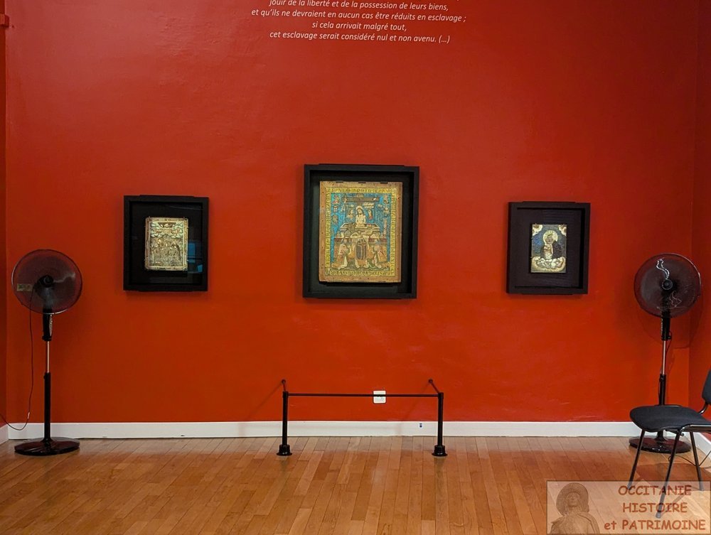 Trois tableaux encadrés sont accrochés sur un mur rouge dans une galerie, encadrés par deux ventilateurs sur pied noirs. Une petite barrière est placée devant, et une chaise est partiellement visible à droite.